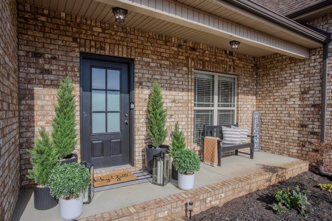 Cozy brick front porch with a black paneled door, potted evergreen plants and lanterns, striped doormat, wooden bench with pillows and a vertical "Welcome" sign.