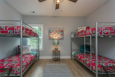 Modern shared kids' bunk room with two metal double bunk beds and bright pink paisley bedding, gray walls, hardwood floors, central rug, round nightstand with lamp and colorful wall art under a ceiling fan.