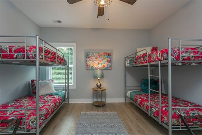 Modern shared kids' bunk room with two metal double bunk beds and bright pink paisley bedding, gray walls, hardwood floors, central rug, round nightstand with lamp and colorful wall art under a ceiling fan.