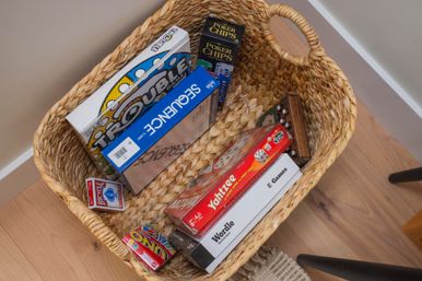 Wicker basket on hardwood floor filled with family game-night favorites — Yahtzee, UNO, Sequence, Trouble, Wordle, poker chips and a deck of playing cards.