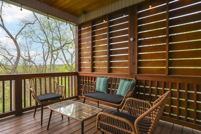 Cozy covered wooden deck with rattan outdoor seating, black cushions and striped pillows, glass-top coffee table, slatted wood privacy wall and treetop view.
