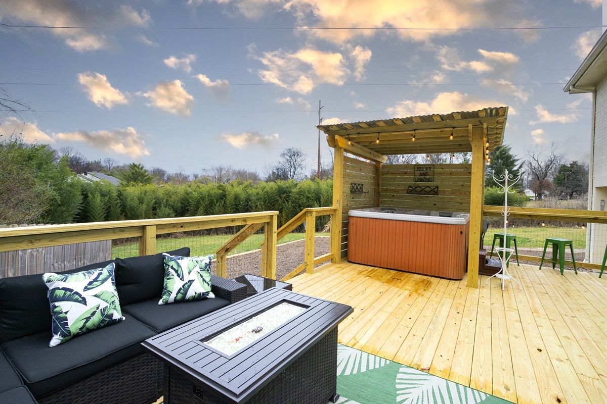 Cozy backyard deck with wooden pergola-covered hot tub, black wicker sofa with tropical pillows, outdoor fire-pit table, string lights and green stools by a pine privacy hedge under a pastel sky