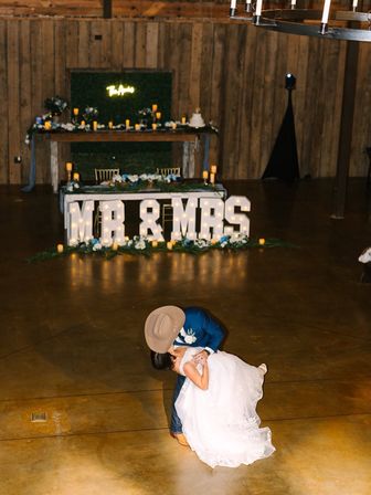 Bride and groom dip during first dance at a rustic barn reception with illuminated "MR & MRS" marquee letters and candlelit greenery