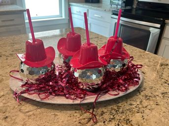 Playful party centerpiece: four silver disco-ball tumblers with bright pink cowboy-hat lids and matching straws, nestled in pink metallic ribbon on a white platter atop a granite kitchen island.