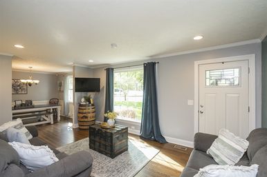 Cozy open-plan living room with gray walls and hardwood floors, sunlight streaming through a large front window with navy curtains, white front door, plush gray sofas, rustic trunk coffee table and barrel TV stand, dining nook visible