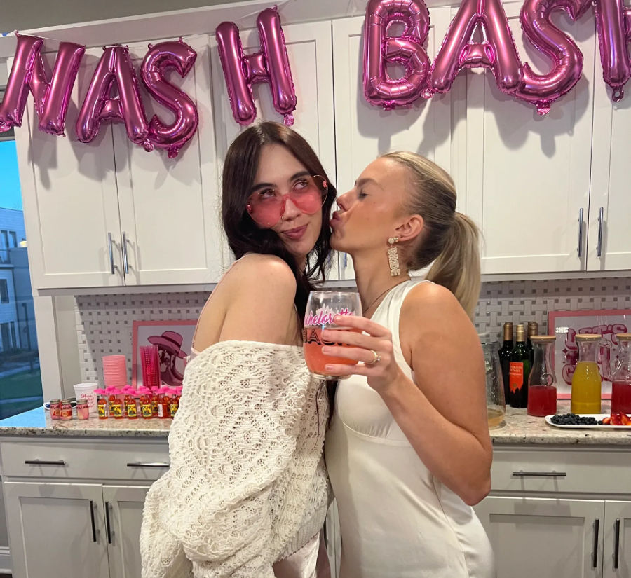 Two friends celebrating in a home kitchen at a pink-themed party — one in a white crochet top with heart sunglasses poses while the other in a white dress kisses her cheek and holds a pink cocktail, foil balloons and a drink spread on the counter.