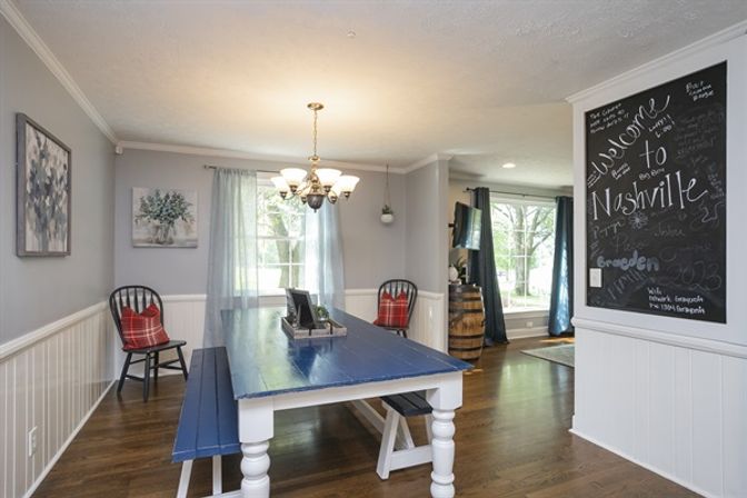 Cheerful farmhouse dining room with a blue plank table and benches, overhead chandelier, chalkboard wall reading "Welcome to Nashville", plaid-accent black chairs, wainscoting and hardwood floors