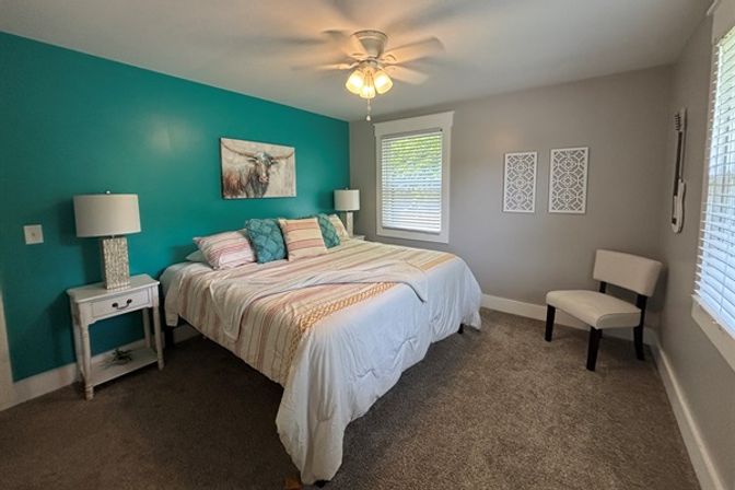 Cozy bedroom with teal accent wall, king-size bed dressed in striped bedding and teal throw pillows, white nightstands and lamps, ceiling fan, gray walls and carpet, and a window with blinds letting in natural light.