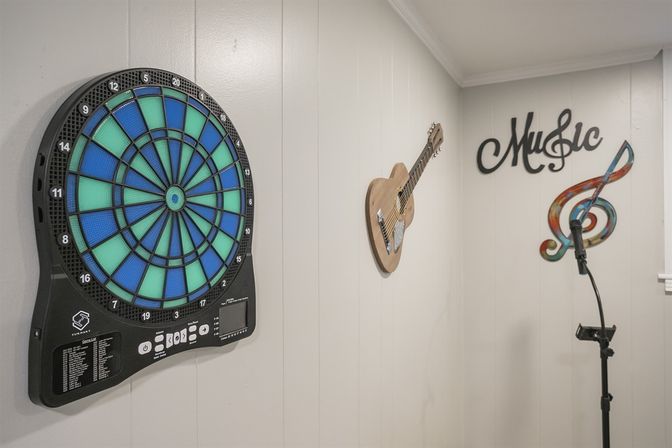 Home music-and-game room corner with blue-green electronic dartboard on white paneled wall, wall-mounted wooden guitar, decorative "Music" sign with colorful treble clef, and a microphone on a stand.