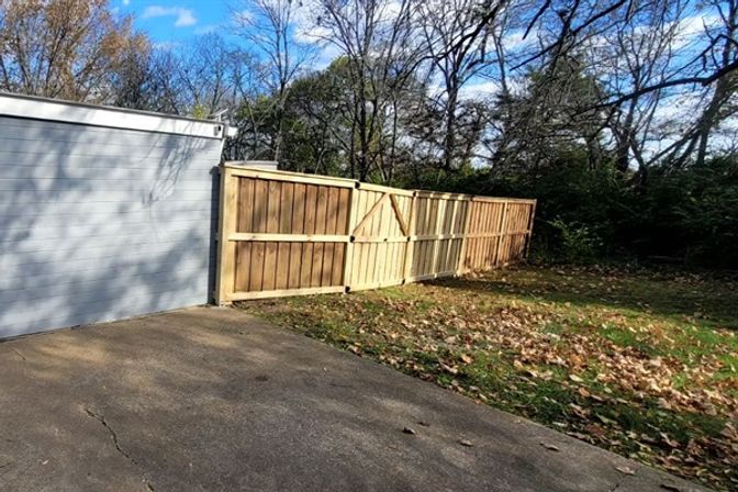 Driveway leading to a light-gray garage and a newly installed wooden privacy fence gate opening to a leaf-strewn suburban backyard with bare trees under a blue autumn sky.