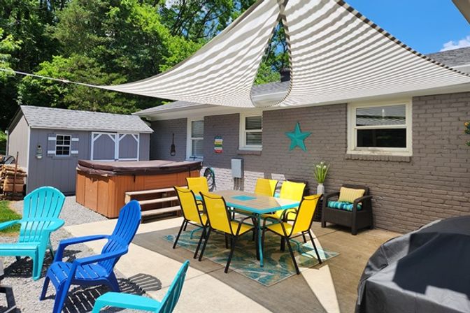 Sunny backyard patio with striped shade sail, yellow outdoor dining set on rug, blue/turquoise Adirondack chairs, hot tub and gray brick house