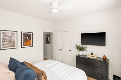 Bright modern minimalist bedroom with white walls and ceiling fan, wall-mounted TV above a black dresser with a potted plant, framed artwork on the wall, and a neatly made bed with blue and brown pillows and sunlight stripes.