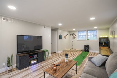 Finished basement home rec room with large flat-screen TV, gray sofa, wood coffee table, shuffleboard, indoor putting green and music corner with microphone and speaker.