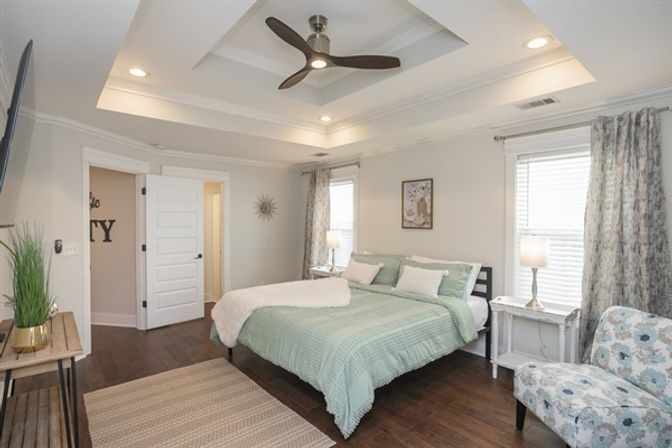 Bright modern master bedroom with tray ceiling and three-blade ceiling fan, dark hardwood floors, mint-green bedding on a queen bed, white nightstands with lamps, patterned accent chair, neutral walls and large windows.