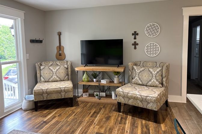 Inviting living room seating nook with two patterned accent chairs, wooden TV console holding a flat-screen, wall-mounted acoustic guitar and decorative crosses, large window with yard view, and warm hardwood floors.