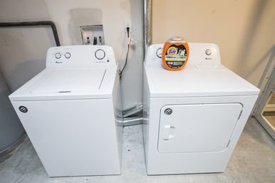 White top-load washer and matching front-load dryer side-by-side in a basement laundry area on a concrete floor, orange detergent container on the dryer, utility hookups and vent duct visible