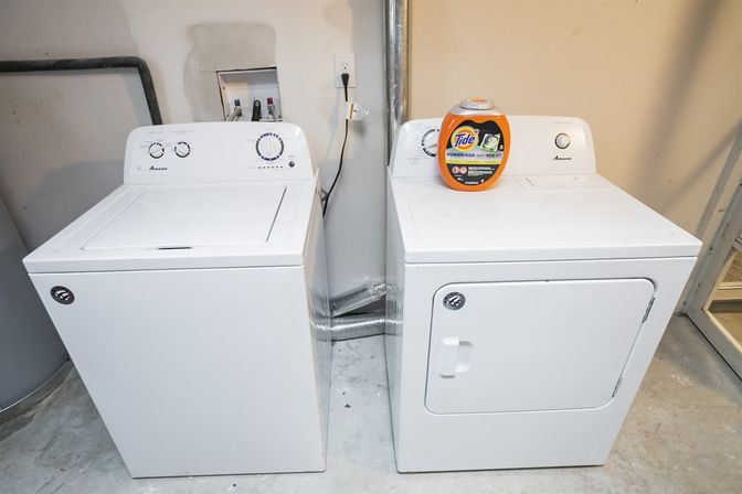 White top-load washer and matching front-load dryer side-by-side in a basement laundry area on a concrete floor, orange detergent container on the dryer, utility hookups and vent duct visible