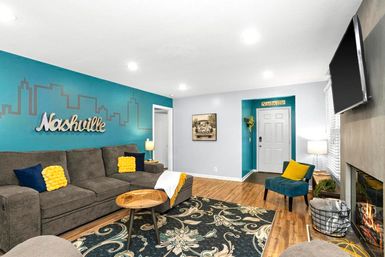 Cozy Nashville-themed living room with teal skyline wall and “Nashville” sign, gray sectional sofa with yellow accent pillows, patterned area rug, wood coffee table, wall-mounted TV and fireplace on hardwood floors.