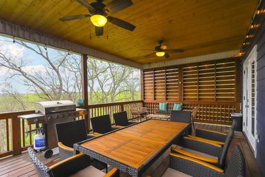 Cozy covered wooden porch with large teak dining table, wicker chairs, ceiling fans, gas grill and lounge seating overlooking a leafy treetop backyard view.