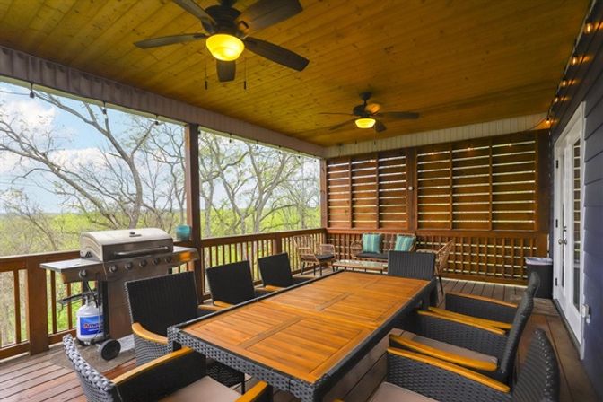 Cozy covered wooden porch with large teak dining table, wicker chairs, ceiling fans, gas grill and lounge seating overlooking a leafy treetop backyard view.
