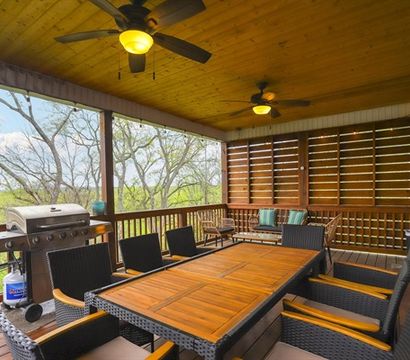 Cozy covered wooden porch with large teak dining table, wicker chairs, ceiling fans, gas grill and lounge seating overlooking a leafy treetop backyard view.