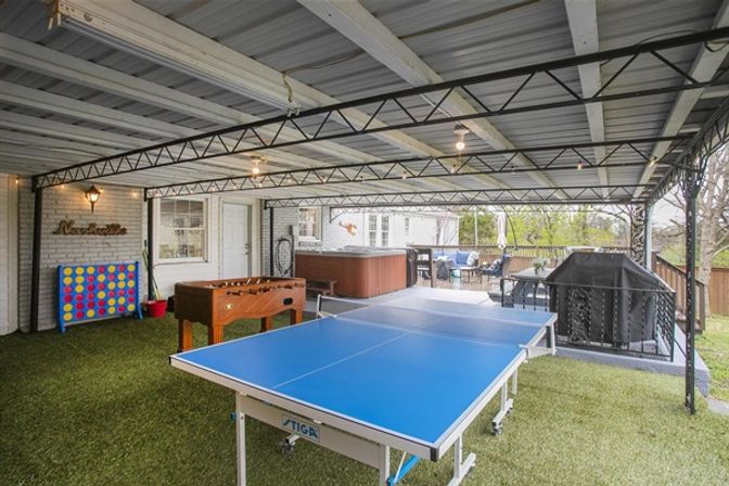 Metal-roofed backyard patio entertainment area with blue ping-pong table, foosball, giant Connect Four, hot tub, grill and lounge seating on artificial turf