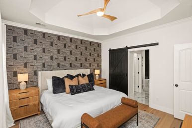 Modern master bedroom with geometric patterned accent wall, white king bed layered with black and tan pillows, wooden nightstands and lamps, brown leather bench, black sliding barn door opening to marble bathroom, tray ceiling with wooden ceiling fan