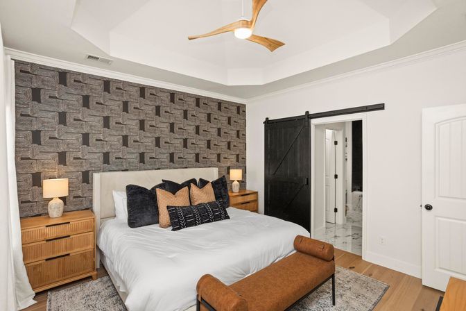 Modern master bedroom with geometric patterned accent wall, white king bed layered with black and tan pillows, wooden nightstands and lamps, brown leather bench, black sliding barn door opening to marble bathroom, tray ceiling with wooden ceiling fan