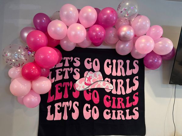Indoor pink balloon garland over a black banner reading LET'S GO GIRLS in bold pink letters with a camo cowgirl hat graphic — fun girls' party decor