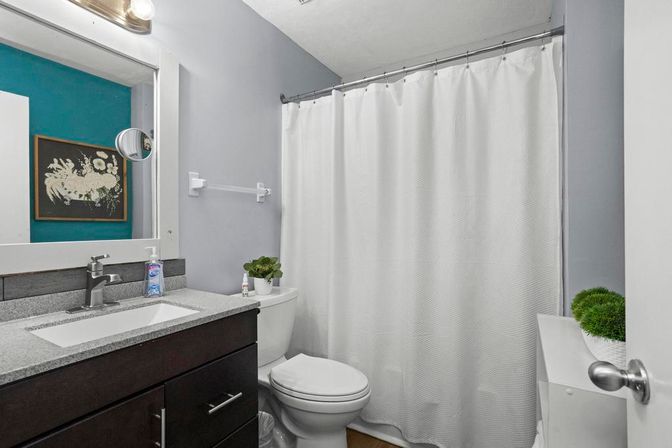 Small modern bathroom with gray walls, white textured shower curtain, dark wood vanity and granite countertop with sink, white toilet, wall mirror and small potted plants for a touch of greenery.