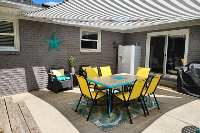 Covered backyard patio with teal-accent tile dining table and six yellow chairs under a striped shade canopy, gray brick wall with teal star, outdoor fridge and grill