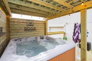 Covered outdoor hot tub on a wooden deck with bubbling water, string lights, a privacy slat fence with a "Relax and Unwind" sign, and striped towels hanging — cozy backyard spa setting.