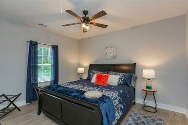 Contemporary bedroom with dark wood sleigh bed dressed in navy floral bedding, fuzzy orange accent pillow, teal glass bedside lamps, ceiling fan, navy curtains, and light hardwood floors.