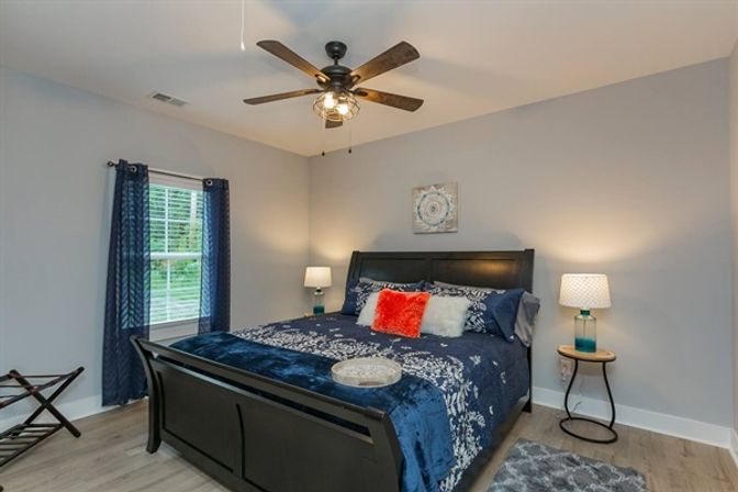 Contemporary bedroom with dark wood sleigh bed dressed in navy floral bedding, fuzzy orange accent pillow, teal glass bedside lamps, ceiling fan, navy curtains, and light hardwood floors.
