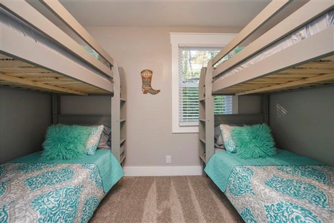 Cozy coastal-inspired bedroom with two gray twin bunk beds facing each other, teal patterned quilts and fluffy teal pillows, white blinds and beige carpet.