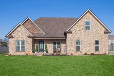 Sunny suburban brick house with brown gabled roof, front porch framed by potted evergreens, multiple large windows with blinds, and a manicured green lawn under a clear blue sky.