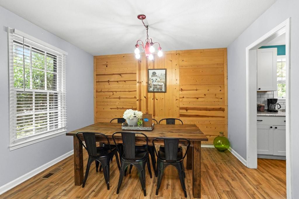 Cozy dining room interior with a rustic wooden accent wall, large farmhouse table with six black metal chairs, hardwood floors, pendant light, window with greenery and open doorway to kitchen.
