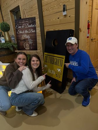 Three smiling adults at a rustic indoor party, pointing at a cornhole board against a wood-paneled wall with a decorative event sign and barrel greenery