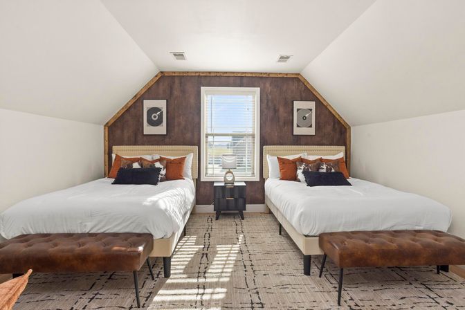 Sunny attic-style bedroom with two identical beds, central window and nightstand, dark wood accent wall, rust throw pillows and leather tufted benches.
