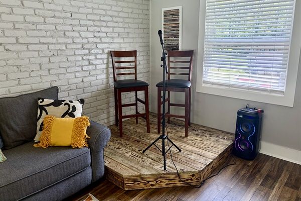 Cozy living room music corner with a small raised wooden stage, two dark wood bar stools and a microphone on a stand; portable speaker with colorful lights by a window with white blinds, gray sofa with decorative pillows against a white brick wall.