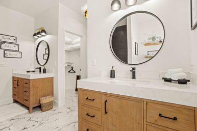 Bright spa-like modern bathroom with two wooden vanities, round black-framed mirrors, white marble countertops and marble tile floor, black faucets, stacked towels and wicker basket.