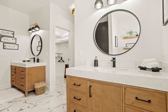 Bright spa-like modern bathroom with two wooden vanities, round black-framed mirrors, white marble countertops and marble tile floor, black faucets, stacked towels and wicker basket.