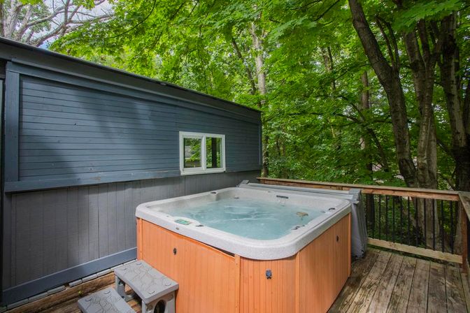 Cozy outdoor hot tub on a wooden deck beside gray siding, surrounded by lush green trees in a private forest setting
