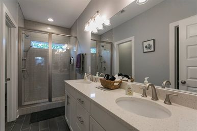 Sleek modern gray master bathroom with double quartz vanity, undermount sinks and brushed-nickel faucets, large mirror, woven basket of towels, and a glass-enclosed walk-in shower with subway tile and dark floor tiles.