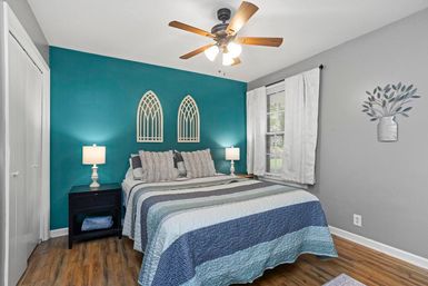 Cozy modern bedroom with teal accent wall, bed dressed in blue-gray striped quilt, matching throw pillows, twin white bedside lamps, arched wall décor, wood ceiling fan and hardwood floors.