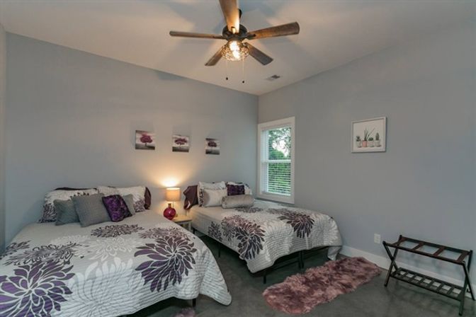 Cozy modern guest bedroom with two purple-and-gray floral beds, neutral gray walls, ceiling fan, nightstand lamp, window, and plush mauve rugs.