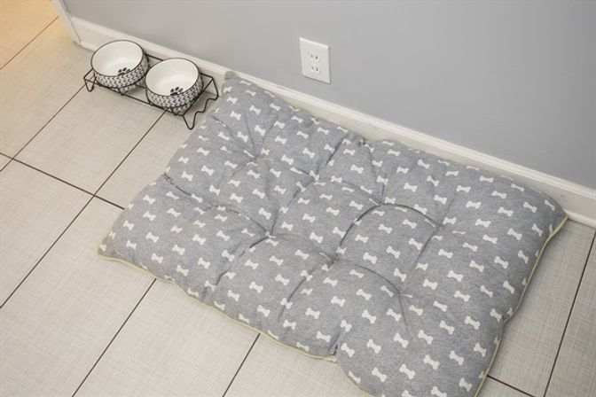 Cozy gray tufted dog bed with white bone pattern on light tiled floor next to black wire elevated double pet bowls against a pale gray wall