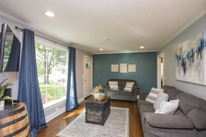 Sunny suburban living room with large picture window and yard view, navy curtains, teal accent wall, gray sofas, rustic trunk coffee table on rug and hardwood floors