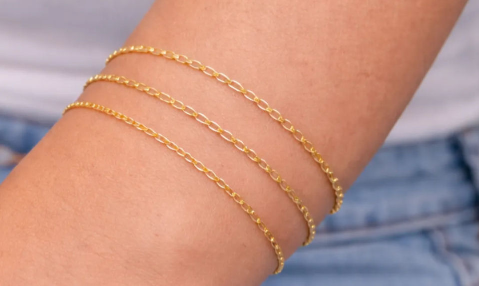 Close-up of forearm wearing three delicate gold chain bracelets with blurred denim jeans and white top in the background.