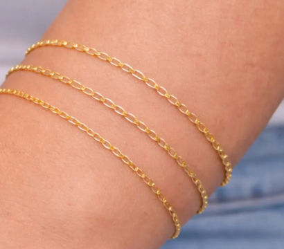 Close-up of forearm wearing three delicate gold chain bracelets with blurred denim jeans and white top in the background.